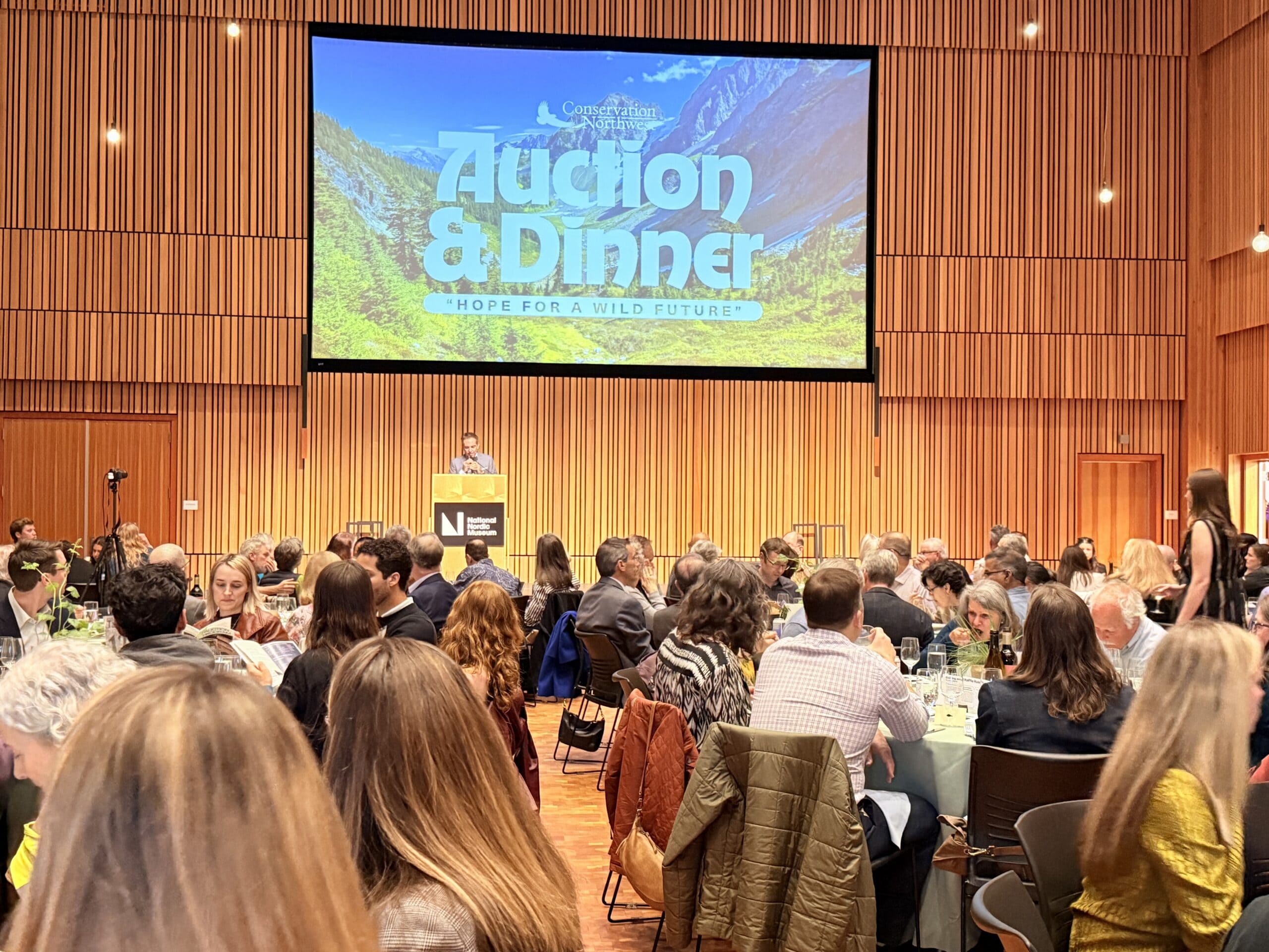 A large group of people sit at tables during an auction and dinner event, with a speaker at a podium and a large screen reading Auction & Dinner: Hope for a Wild Future in the background.